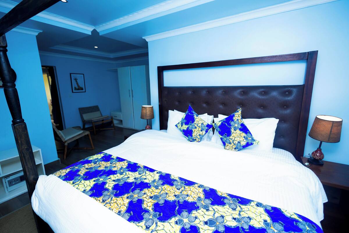 Una cama grande con cabecera capitonada, una colcha de cuadros azules y amarillos, y almohadas a juego. Hay dos lámparas de noche y una silla en la habitación.