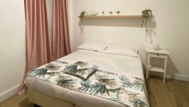 Ein Schlafzimmer mit einem Bett, einem Nachttisch, einer Pflanzenablage und einem Fenster mit Vorhängen.