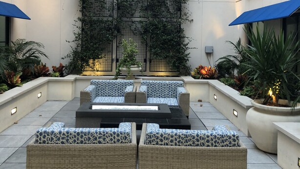 An outdoor seating area with wicker furniture and blue umbrellas, surrounded by potted plants and a decorative wall with climbing plants.