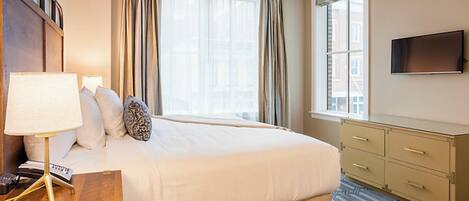 A hotel room with a large bed, a desk with a lamp, a flat-screen TV, and a window with curtains.