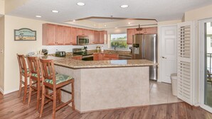 A kitchen with wooden cabinets, a stainless steel refrigerator, a microwave, and a granite countertop. - Oceanwalk 3BR Corner Condo | Private Balcony & 4-Min Walk to Beach (New Smyrna Beach)