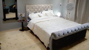 Un dormitorio con una cama y cabecera capitonada, una mesita de noche con lámpara, un espejo y una lámpara de pie.