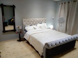 Quarto com cama e cabeceira estofada, mesa de cabeceira com abajur, espelho e luminária de chão.