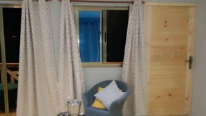 Una habitación con puerta de madera, una silla con cojín, una mesita con vela y una ventana con cortinas.