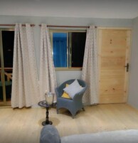 Um quarto com porta de madeira, uma cadeira com almofada, uma pequena mesa com vela e uma janela com cortinas.