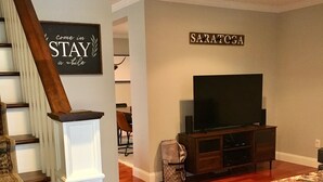 Un salon avec un escalier, un téléviseur à écran plat et une enseigne sur laquelle on peut lire « SARATOGA ».