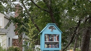A quaint blue library house with a white picket fence, surrounded by lush greenery and trees. - Breezy Vibes Cottage (3bd) ~ Backyard Blissful  ~ Walk to beaches (South Yarmouth)