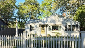 A single-story house with a white picket fence, a front porch, and a small garden area with potted plants. - Breezy Vibes Cottage (3bd) ~ Backyard Blissful  ~ Walk to beaches (South Yarmouth)