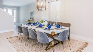 Een eetruimte met een lange tafel gedekt voor zes personen, met grijs gestoffeerde stoelen met gouden poten, een kamerbreed tapijt en een geometrisch hanglamp.
