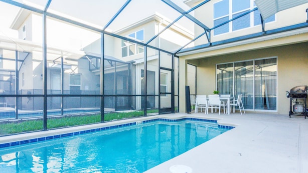 An indoor swimming pool with clear glass walls, a white tiled floor, and a seating area with a table and chairs.