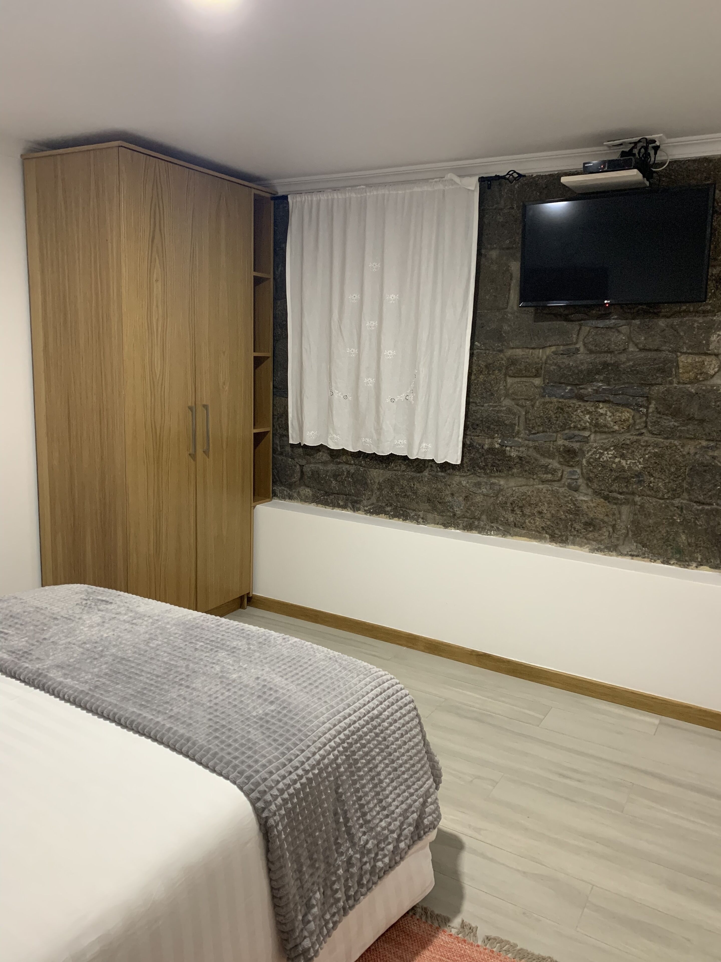 A bedroom with a bed, a wardrobe, a stone wall, a TV mounted on the wall, and a window with curtains.