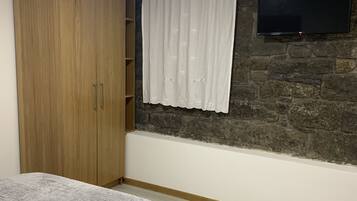 A bedroom with a bed, a wardrobe, a stone wall, a TV mounted on the wall, and a window with curtains.