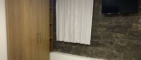 A bedroom with a bed, a wardrobe, a stone wall, a TV mounted on the wall, and a window with curtains.