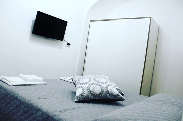 A hotel room with a bed, a grey blanket, a white pillow with a circular pattern, a folded white towel, a flat-screen TV mounted on the wall, and a white wardrobe.