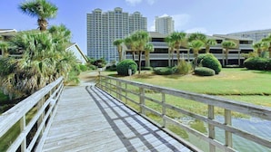 A wooden boardwalk leading to a building complex with multiple floors and a grassy area with palm trees. - Seascape Amenities☀️Multiple Community Pools☀️Walk to Beach-Casa Bellal⛱️ (Miramar Beach)