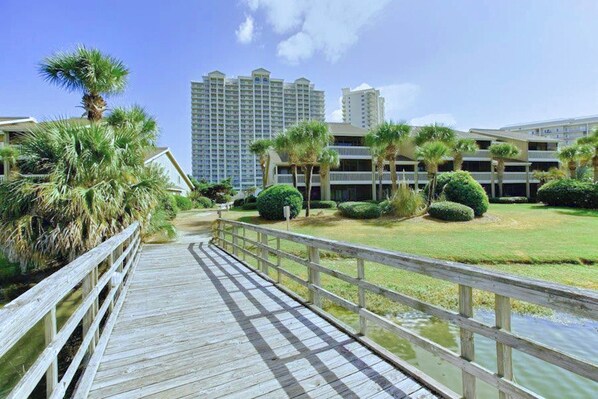 A wooden boardwalk leading to a building complex with multiple floors and a grassy area with palm trees.