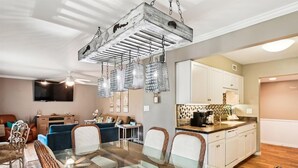 A modern living room with a glass dining table, blue sofas, and a kitchen area with white cabinets and a tiled backsplash. - Seascape Amenities☀️Multiple Community Pools☀️Walk to Beach-Casa Bellal⛱️ (Miramar Beach)