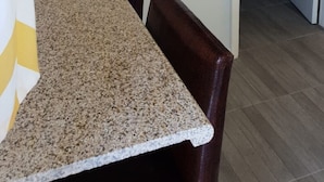 Una habitación con encimera de granito, una mesa de comedor, sillas de madera oscura, un televisor de pantalla plana montado en la pared y una puerta que da a otra habitación.