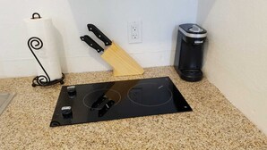 Una cocina con tope de inducción negro, tabla de cortar de madera, un rollo de toallas de papel y un cubo de basura negro.