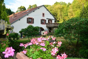 Uma casa branca com telhado de telhas vermelhas, um deck de madeira e um jardim com flores cor-de-rosa.