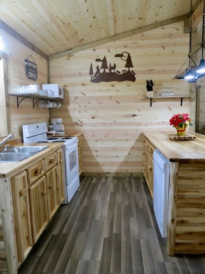 Une cuisine avec des meubles de rangement en bois, une cuisinière, un four à micro-ondes et une machine à café. Il y a un panneau accroché au mur et une étagère avec des tasses et des verres.