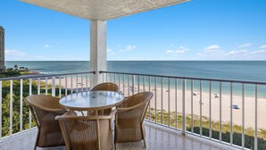 Un balcon avec des fauteuils en osier et une table en verre, donnant sur une plage où l’on aperçoit des gens et des parasols.