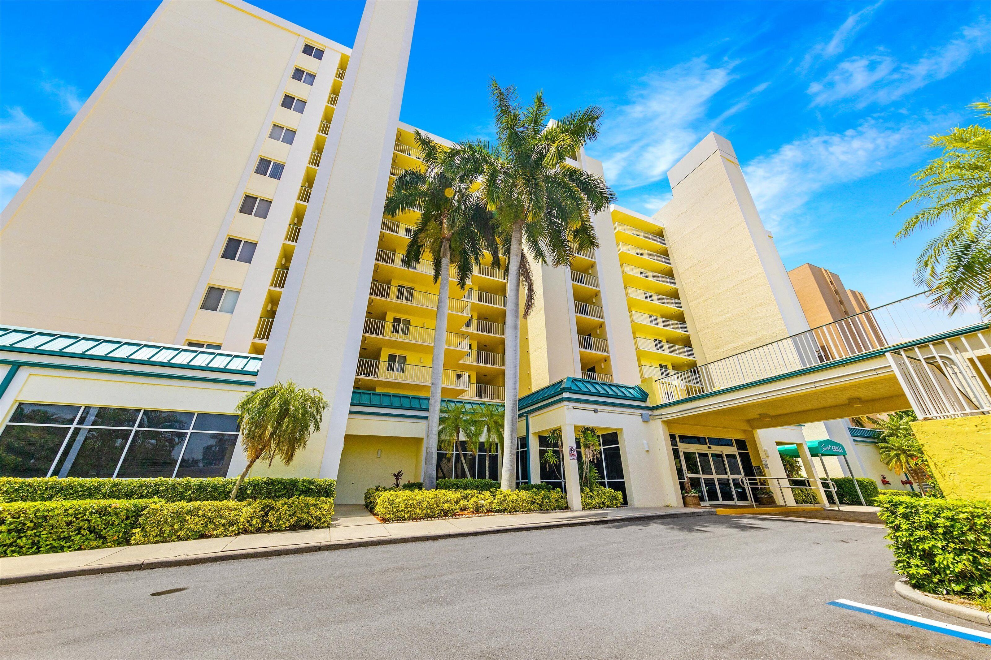 Un hotel de varios pisos con un atrio central, palmeras y un cielo azul despejado.