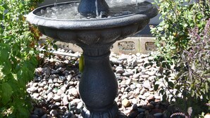 Ett fontänkomplex i flera nivåer, byggt av sten, med vatten som forsar ner, omgivet av stenar och blommande växter.