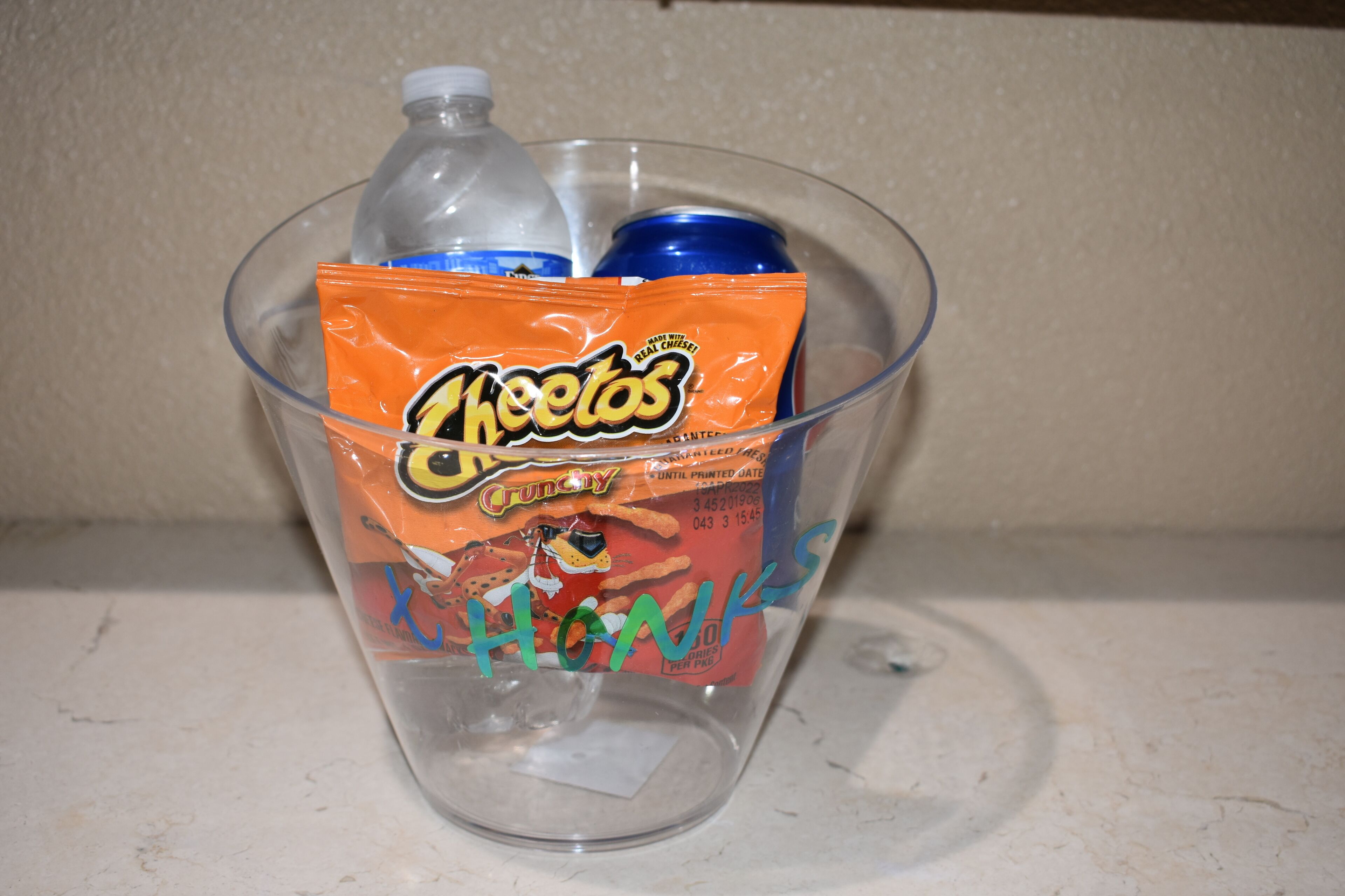 A clear plastic cup containing a bag of Cheetos, a bottle of water, and a can of soda.