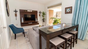 Ein modernes Wohnzimmer mit einer Couch, einem blauen Sessel, einem Glastisch mit Barhocker und einem Wand-Fernseher, dazu eine Zimmerpflanze.
