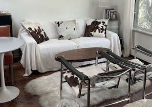 A white sofa with cowhide pillows, a round wooden table, and a metal chair with a fur rug.