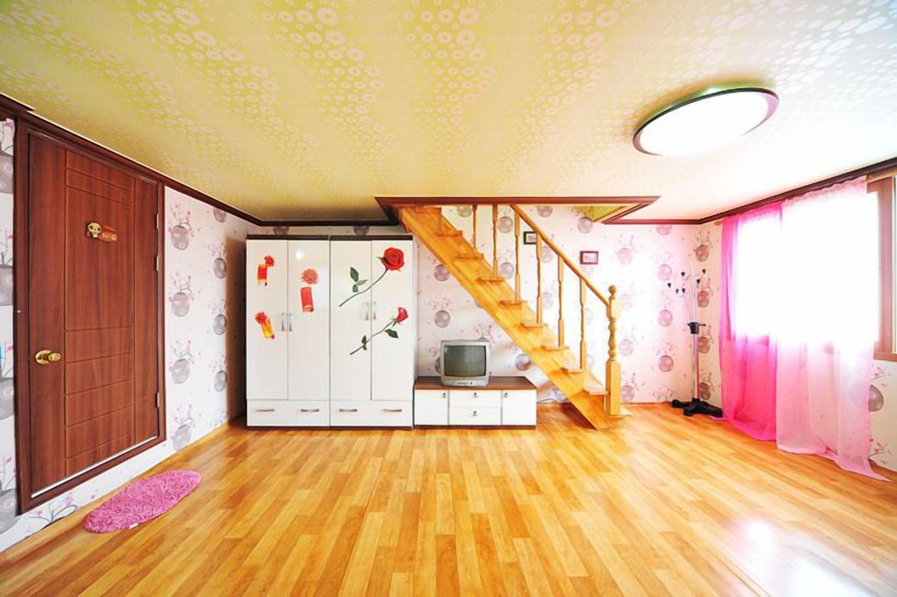 Ein Zimmer mit Holzboden, einer Holztreppe, einem Kleiderschrank, einem Fernseher und einem rosa Vorhang.