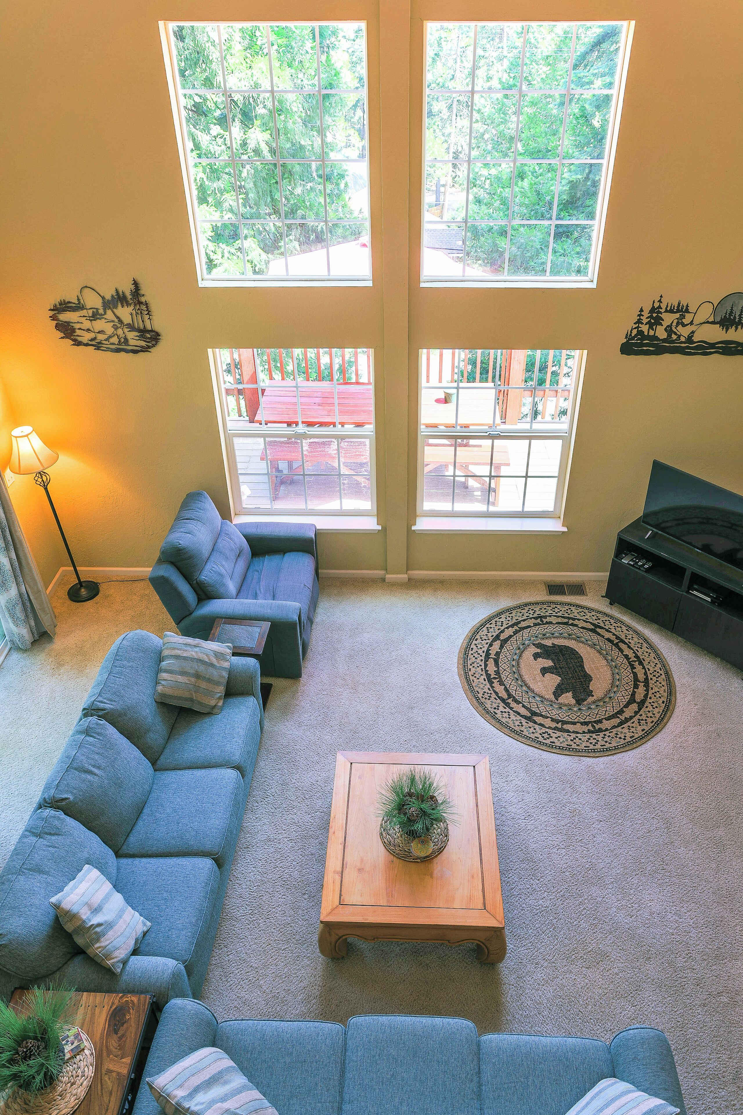 Uma sala de estar com sofá seccional, uma mesa de centro com uma planta e um tapete com estampa de urso. Há duas janelas com vista para o verde lá fora.