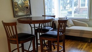 A dining area with a wooden table and four chairs, a sofa, and a window with blinds.