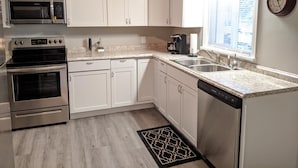 A modern kitchen with white cabinets, stainless steel appliances, a window with blinds, and a clock on the wall.