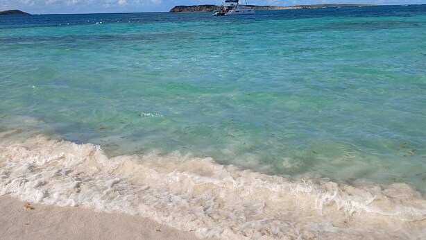 Une plage à l’eau turquoise et limpide, un voilier à l’horizon et une rive sablonneuse.