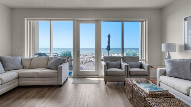 A spacious living room with a large sliding glass door leading to a balcony with a view of the ocean. There are two sofas and a coffee table with a tray of magazines.