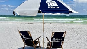 Two beach chairs with 'The Perch' branding under a beach umbrella on a sandy beach with the ocean in the background.