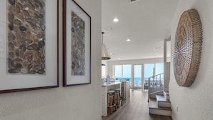 A modern kitchen with a view to the ocean, a staircase leading to a balcony, and framed artwork on the walls.
