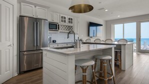 A modern kitchen with stainless steel appliances, a large island with stools, and a view of the ocean through large windows.