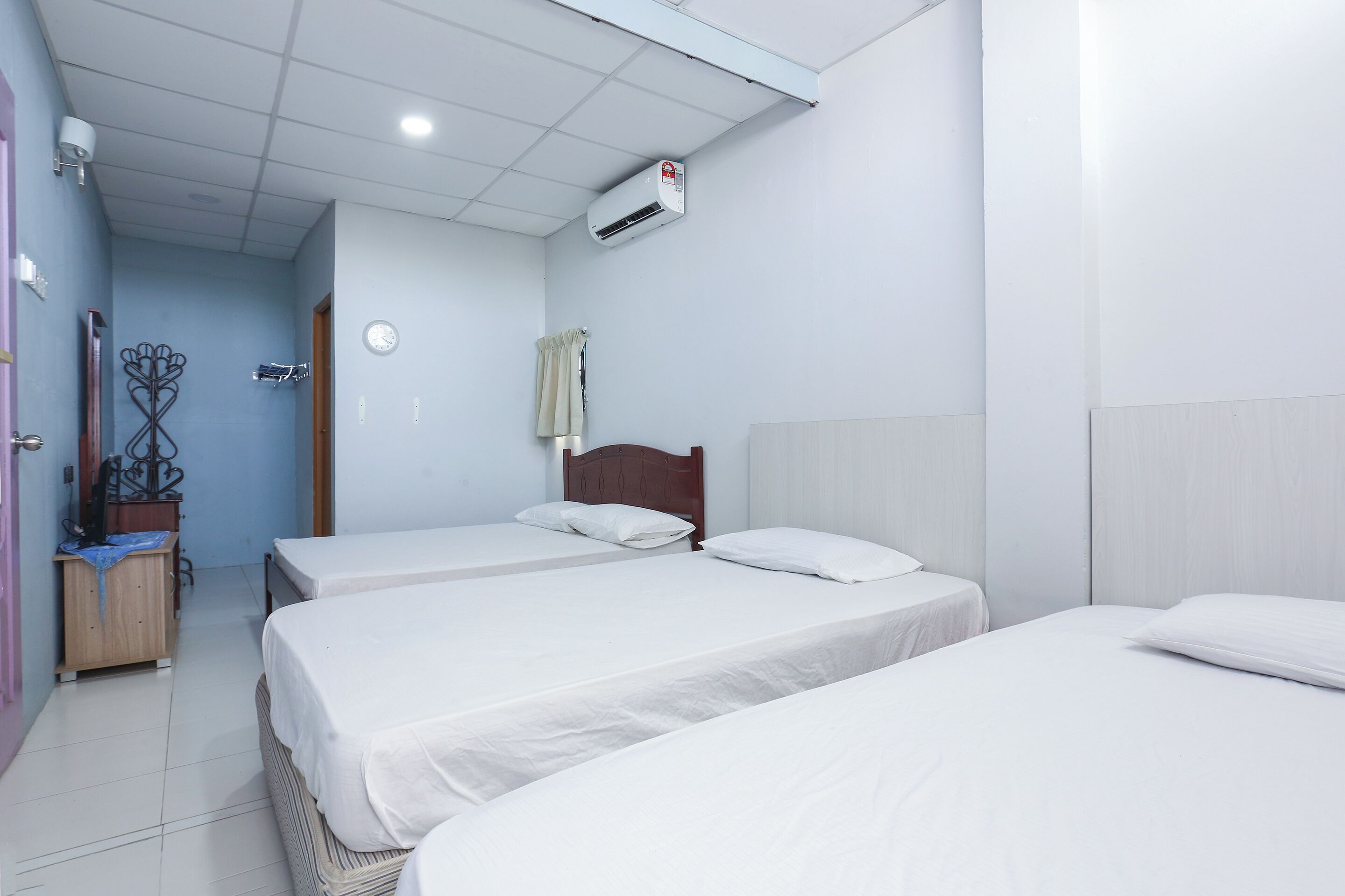 A hotel room with three single beds, a wooden wardrobe, a desk with a chair, a wall-mounted air conditioner, and a clock.