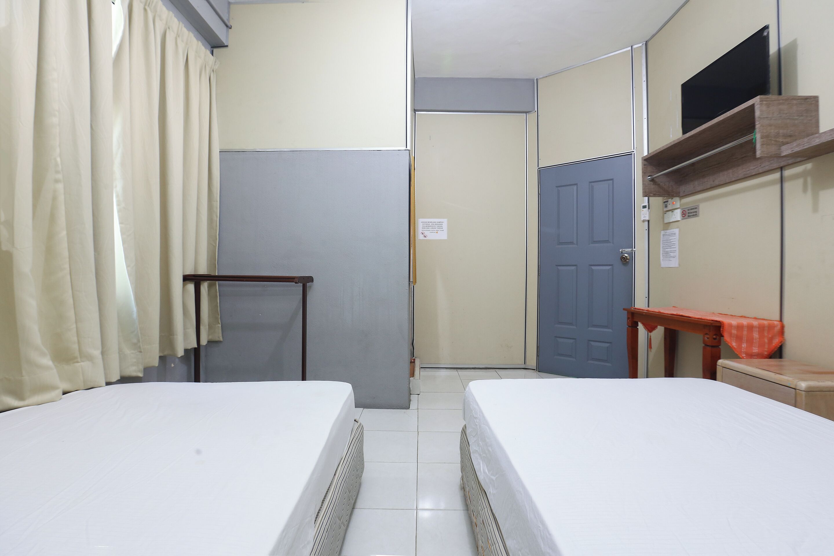 A small, clean room with two single beds, a wooden table, a blue door, and a small shelf.