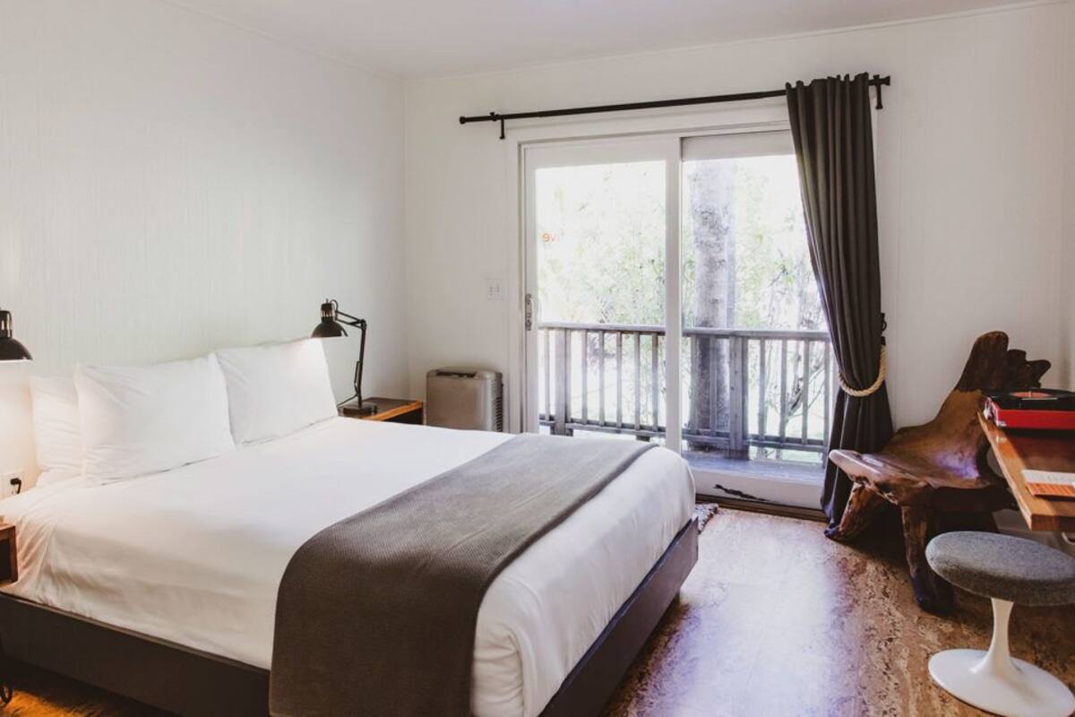 A bedroom with a bed, bedside lamps, a desk with a chair, and a view of the outdoors through a sliding door.