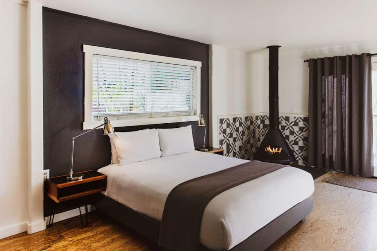 A modern bedroom with a large bed, a window with blinds, a nightstand, a lamp, and a decorative wall panel.