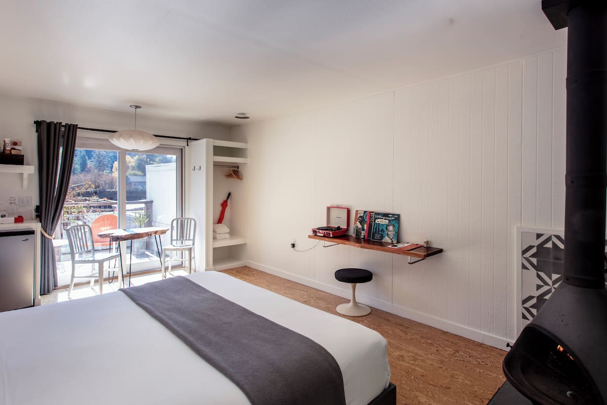 A modern hotel room with a bed, a small table, a chair, a shelf with books, a red umbrella, and a black stove.