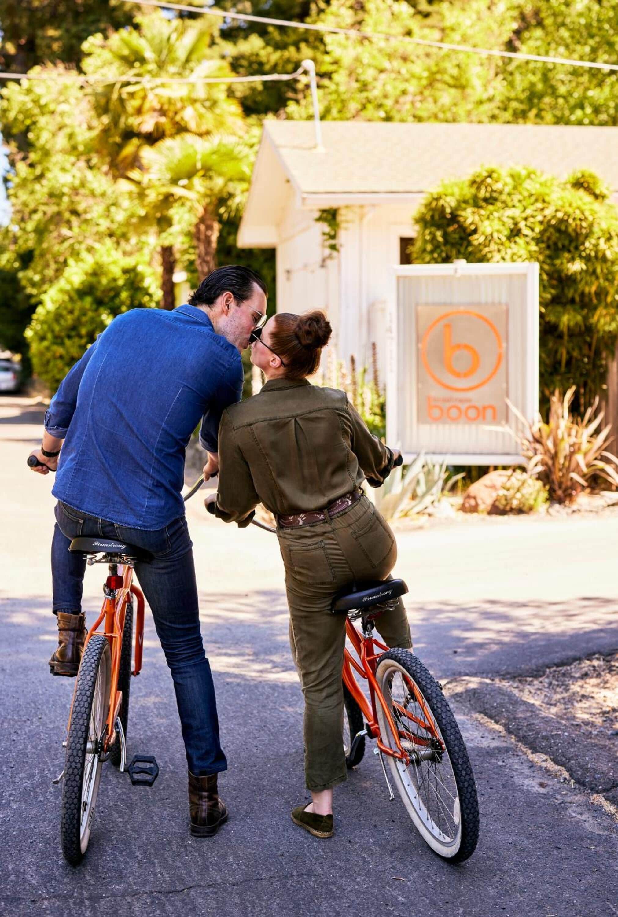 Dos personas en bicicletas, una sosteniendo a la otra, frente a un edificio con un letrero que dice 'boon'.