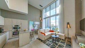 Une cuisine moderne dotée d’appareils électroménagers intégrés, un coin repas avec une table et des chaises blanches, un salon avec un canapé et une table basse, et de grandes fenêtres offrant une vue sur la ville.