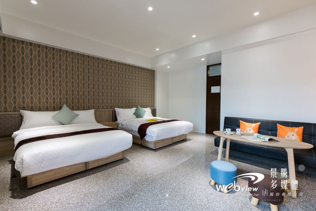 A modern hotel room with two beds, a sofa, a small table, and a patterned wall.