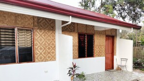Une maison avec un toit rouge, une porte en bois et une fenêtre ornée d’un volet à motifs. Deux plantes en pot sont posées sur le porche.