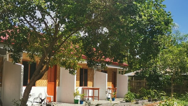 Une maison à deux étages, avec un toit rouge, entourée d’un jardin où poussent différentes espèces de plantes et bordée d’un chemin de gravier.